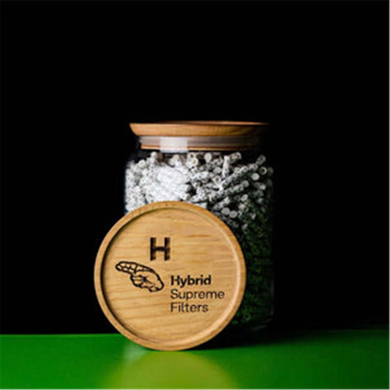 Hybrid Supreme Filters 1000er Glasdose geschlossen – zylindrisches Glasgefäß gefüllt mit weiß-grauen Filtern, mit Holzdeckel mit eingeprägtem Hybrid Supreme Filters Logo, davor liegender Holzdeckel, grüne Fläche, schwarzer Hintergrund